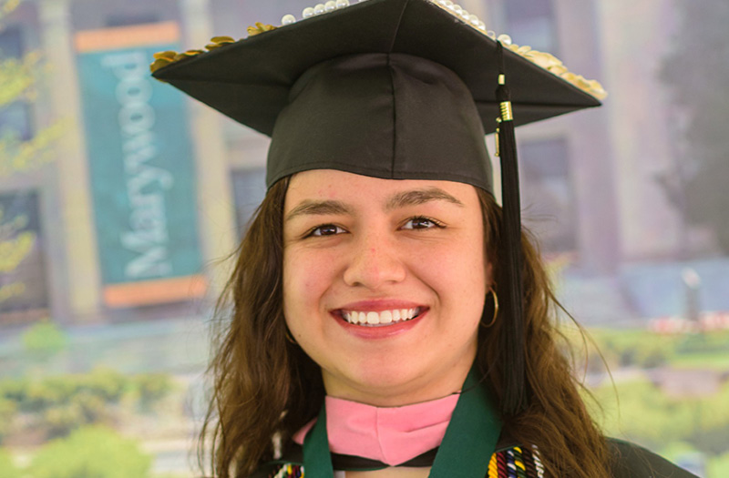 A woman wearing a commencement cap and gown with Marywood University in the background.